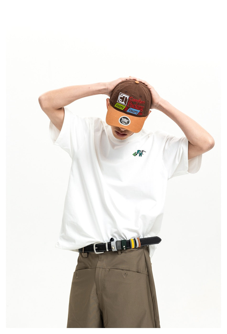 Model wearing a white oversized T-shirt with a small colorful chest graphic, brown trousers, and a brown-and-orange cap.