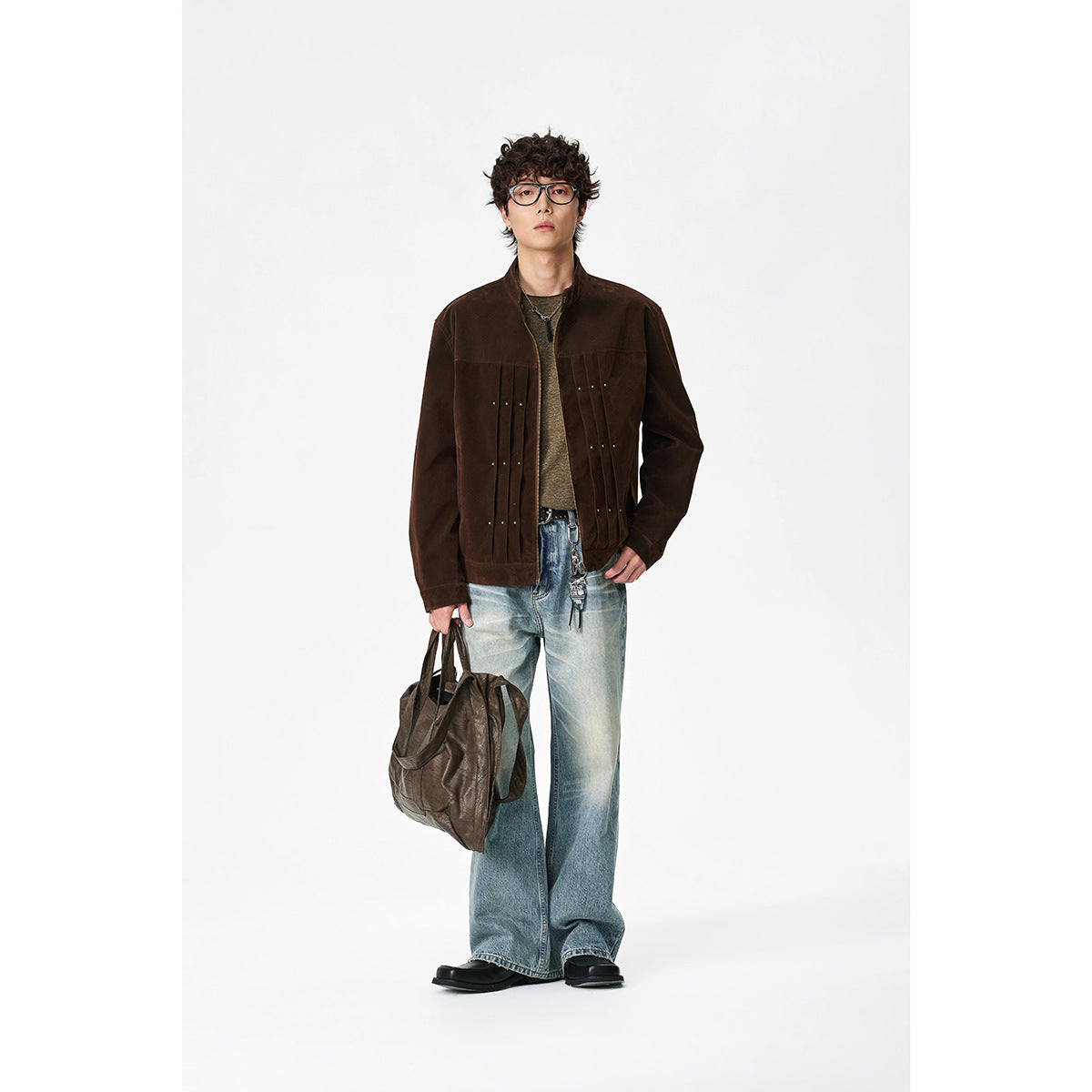 Model wearing a dark brown zip-front jacket with pleated panels and brass studs, styled with jeans and a shoulder bag.