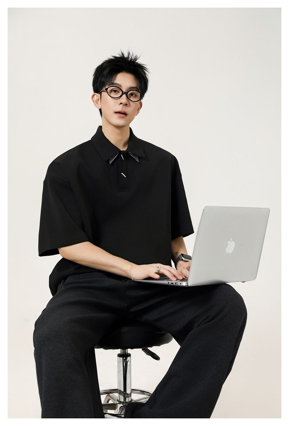 Seated model wearing a black oversized short-sleeve polo with a pointed collar and small decorative accents.