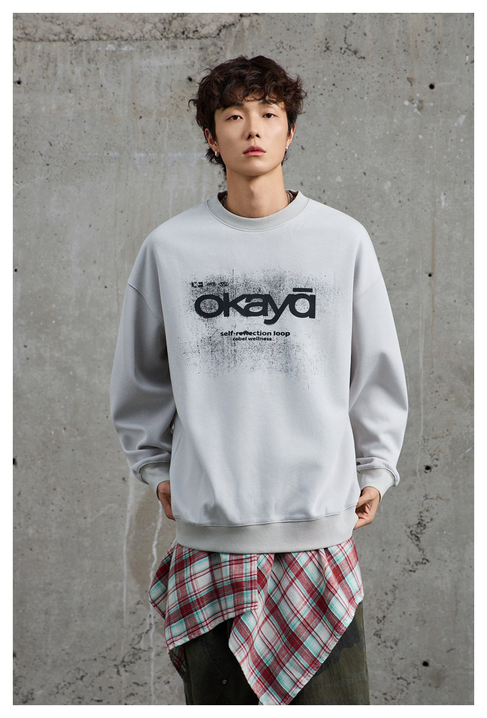 Front view of a model wearing a light gray sweatshirt with a black graphic and a plaid shirt tied at the waist.