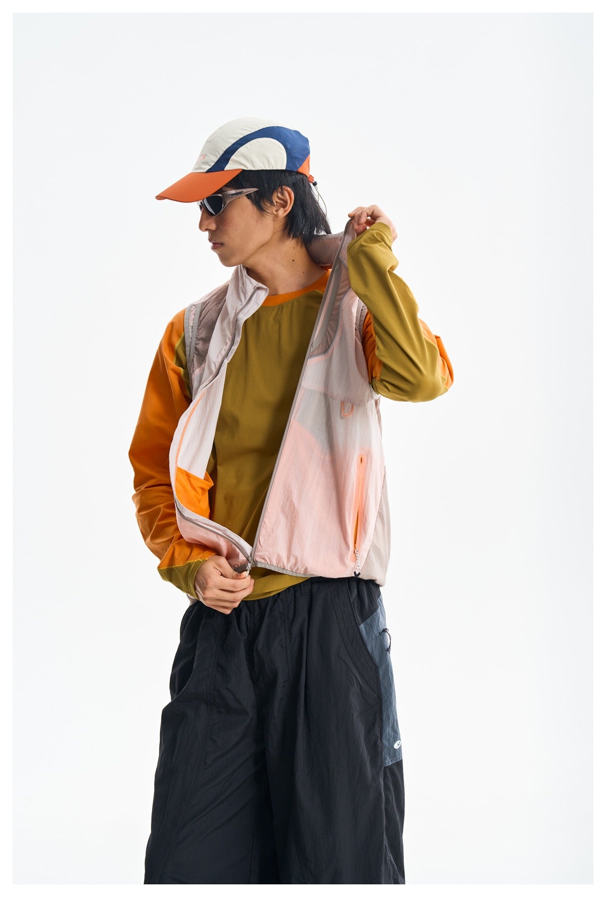 Side view of a model opening a pale pink zip-front vest over an orange and olive top; black pants are also visible.