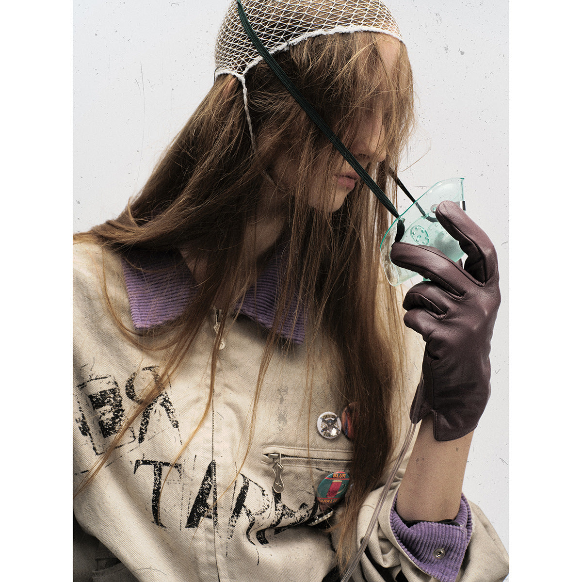 Close-up of a model wearing a distressed beige jacket with a purple collar, graphic print, pins and dark gloves.