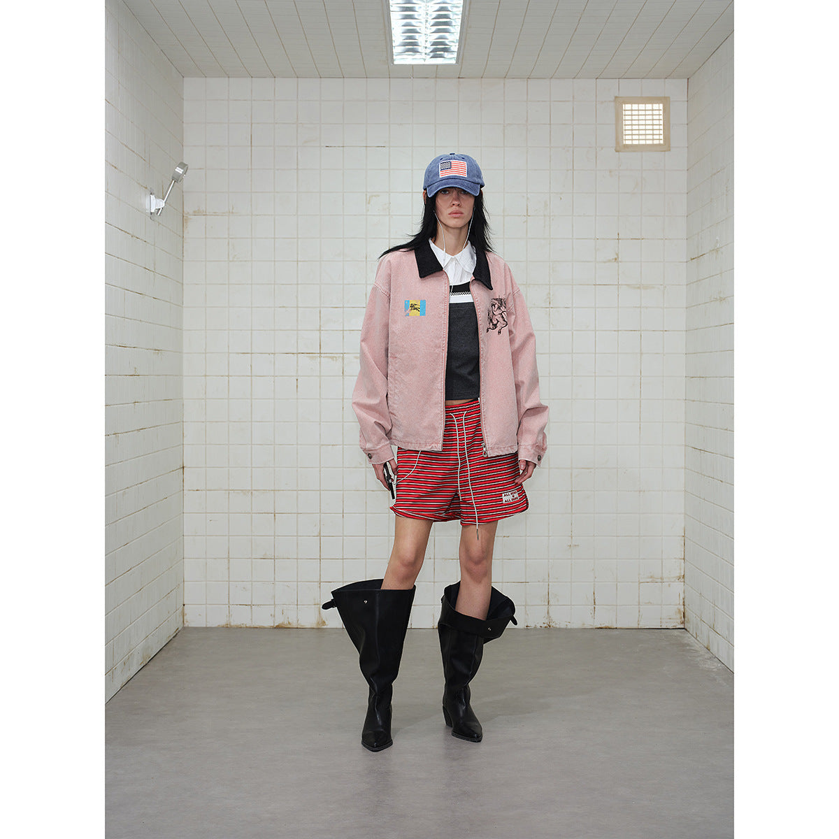 Front-facing model in a dusty pink graphic jacket with black collar, red striped shorts, blue cap, and black boots.