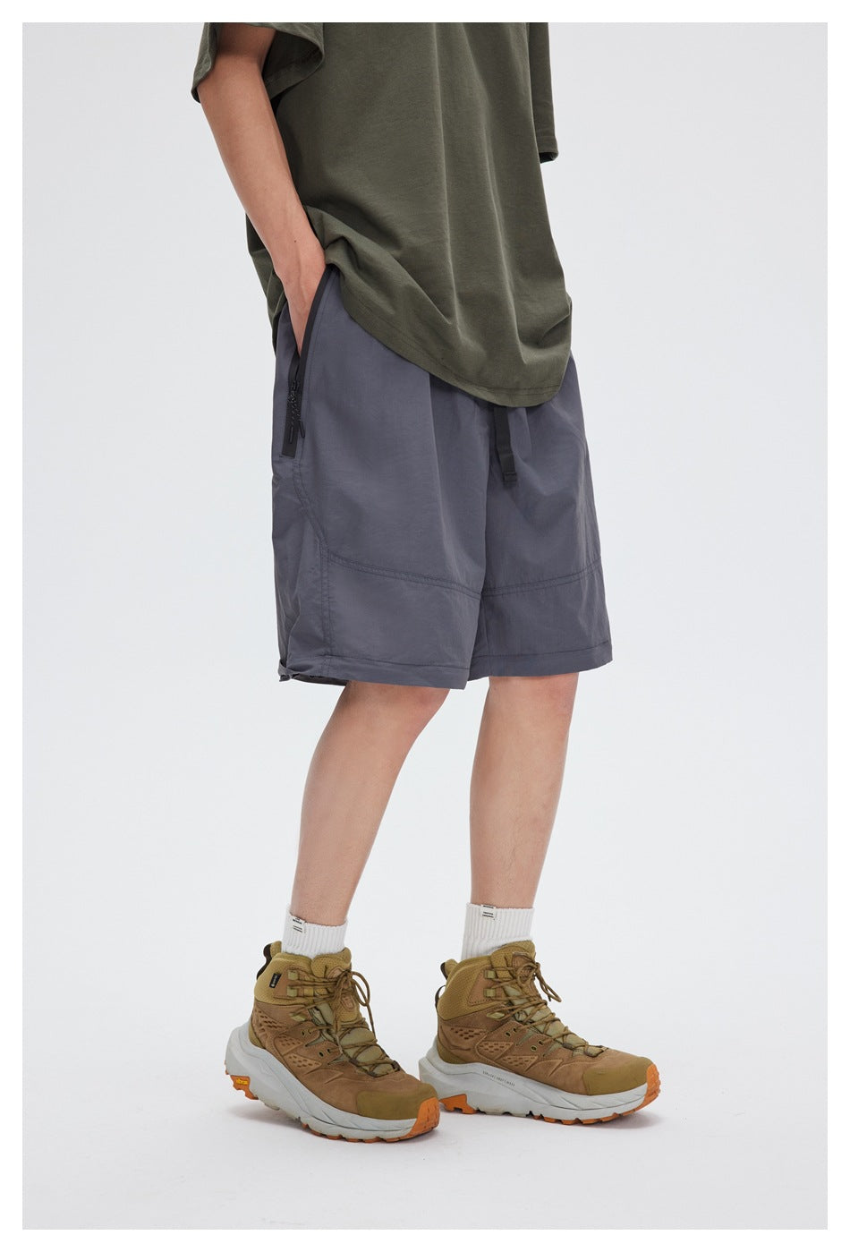 Model wearing loose slate-gray shorts with black zip pockets and a long olive T-shirt, shown at an angle.