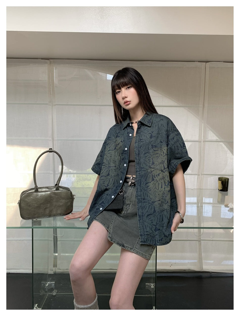 Model wearing an oversized blue-gray botanical-print short-sleeve button-up shirt over a top and denim skirt.
