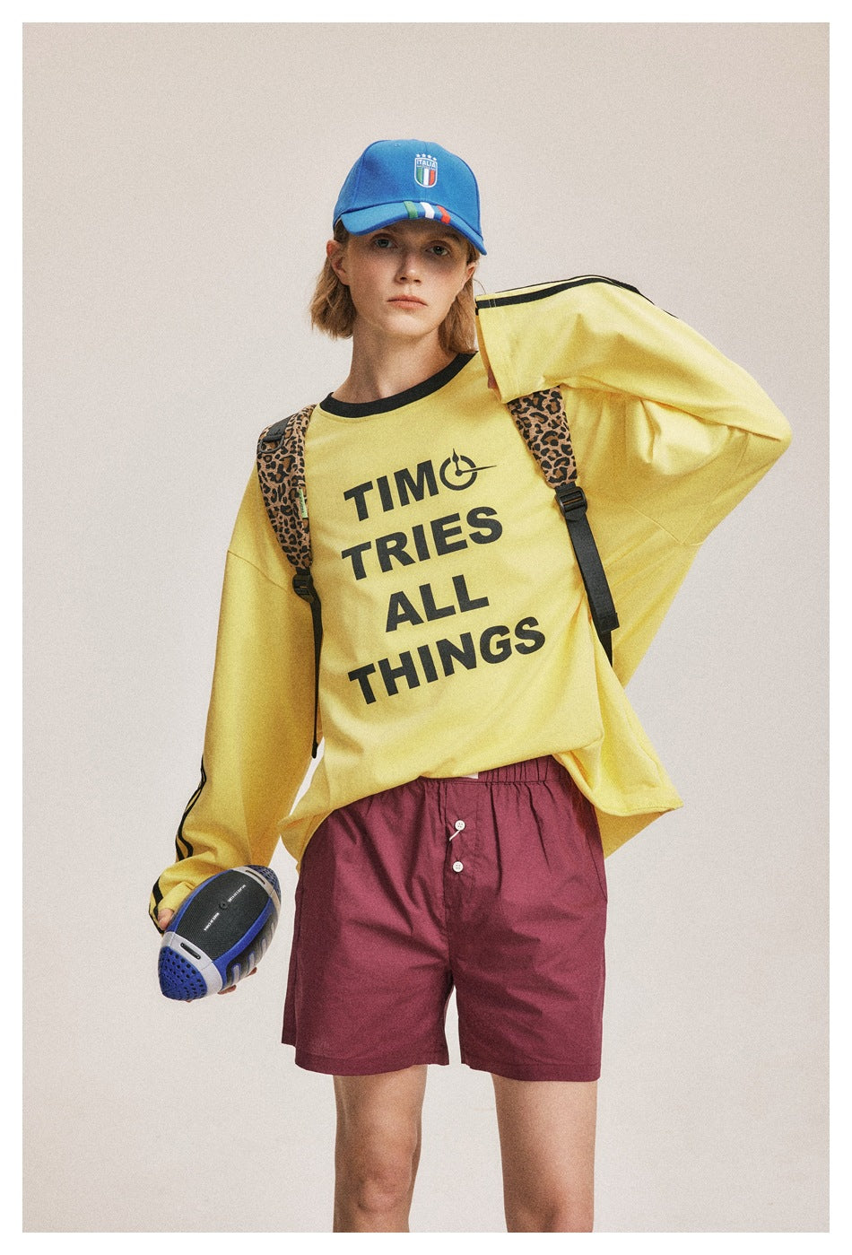 Model wearing a yellow slogan sweatshirt with black sleeve stripes and burgundy shorts, holding a ball.