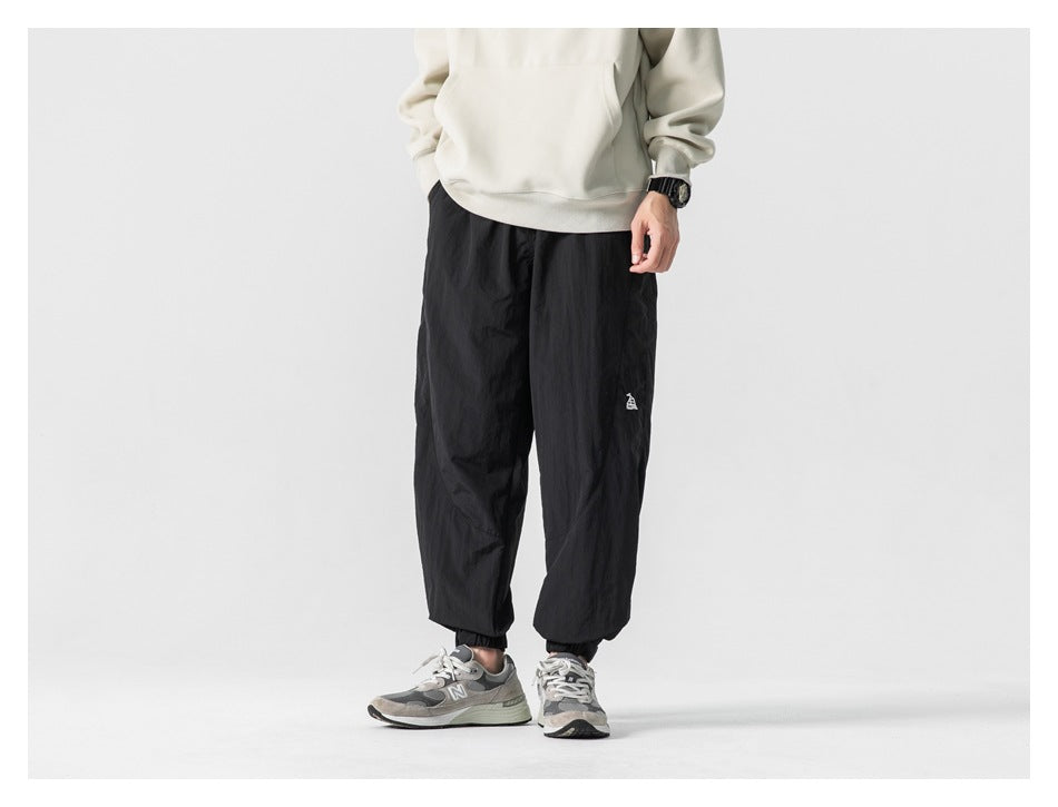 Cropped model wearing loose black jogger pants with elastic cuffs and a small white thigh emblem, shown from the front.