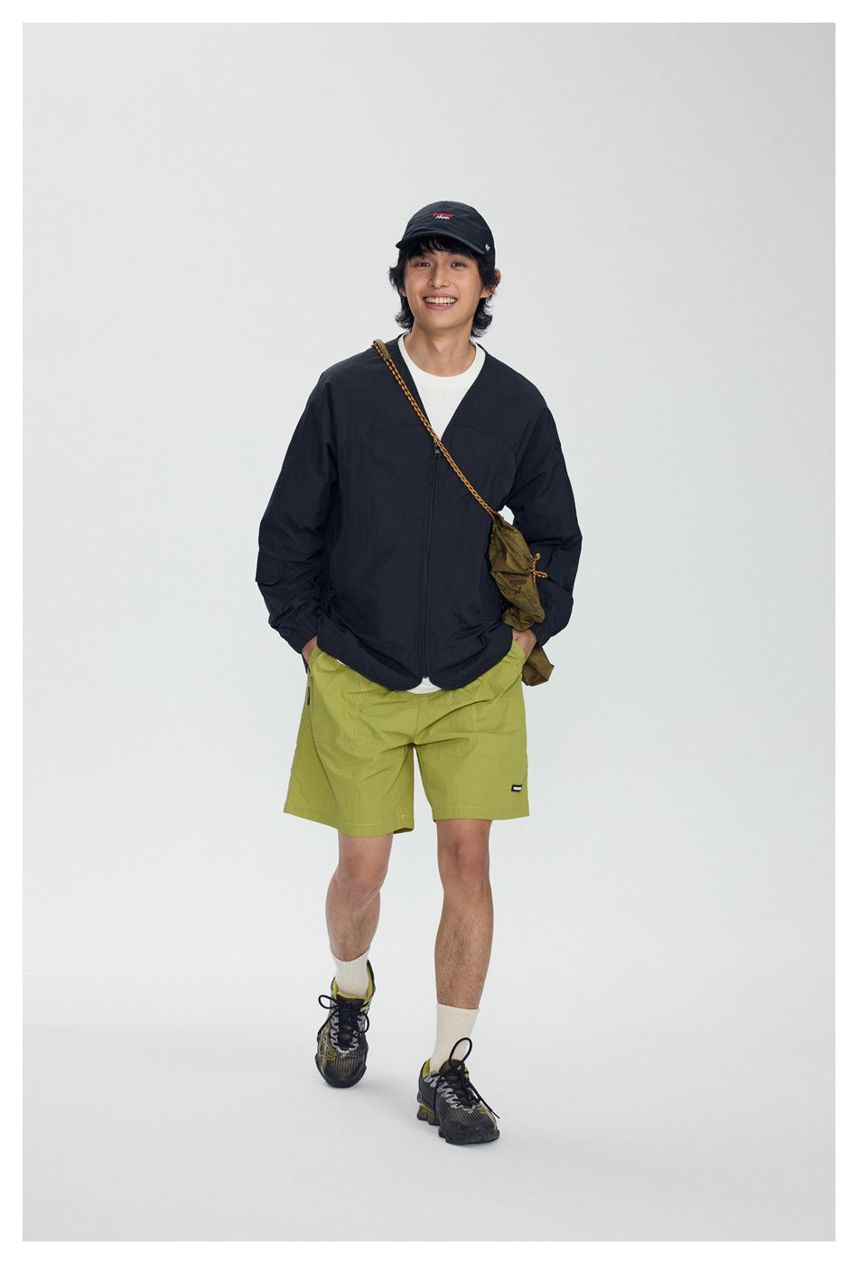 Full-length model wearing a black zip-front jacket, white T-shirt, lime-green shorts, and a crossbody bag.