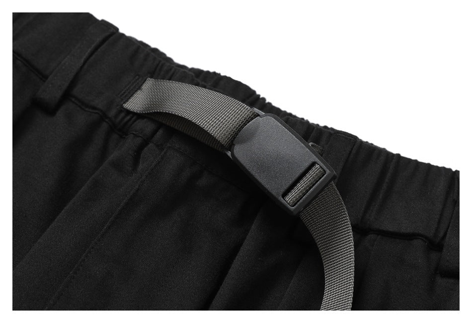 Close-up of black trousers showing the gathered waistband and adjustable webbing belt with a plastic buckle.
