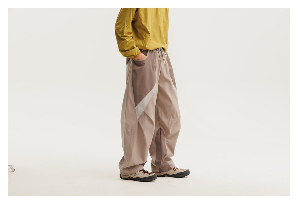 Model wearing loose taupe trousers with dark panels and a pale curved side stripe, shown from a side-front angle.