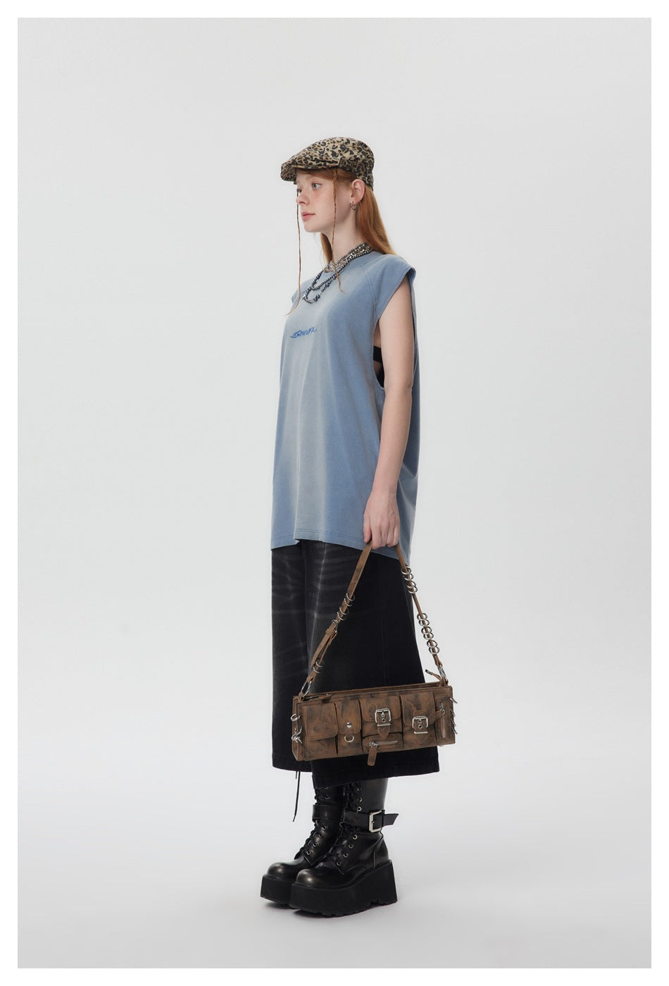 Model in a blue sleeveless top with a leopard cap, dark wide-leg shorts, boots and a brown shoulder bag, side view