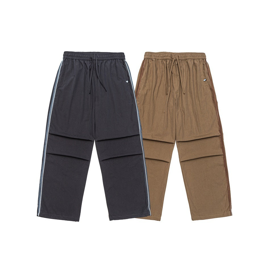 Two pairs of loose trousers shown flat against white: charcoal with pale piping and tan with darker piping.