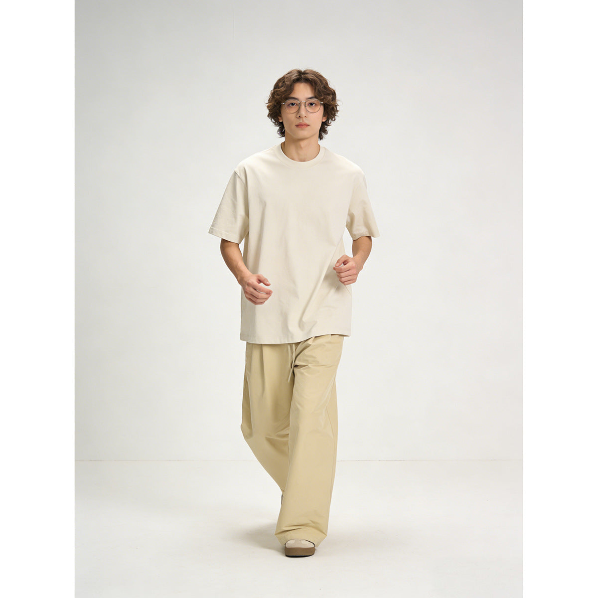 Model wearing an oversized cream T-shirt with loose tan trousers, shown walking against a plain studio background.