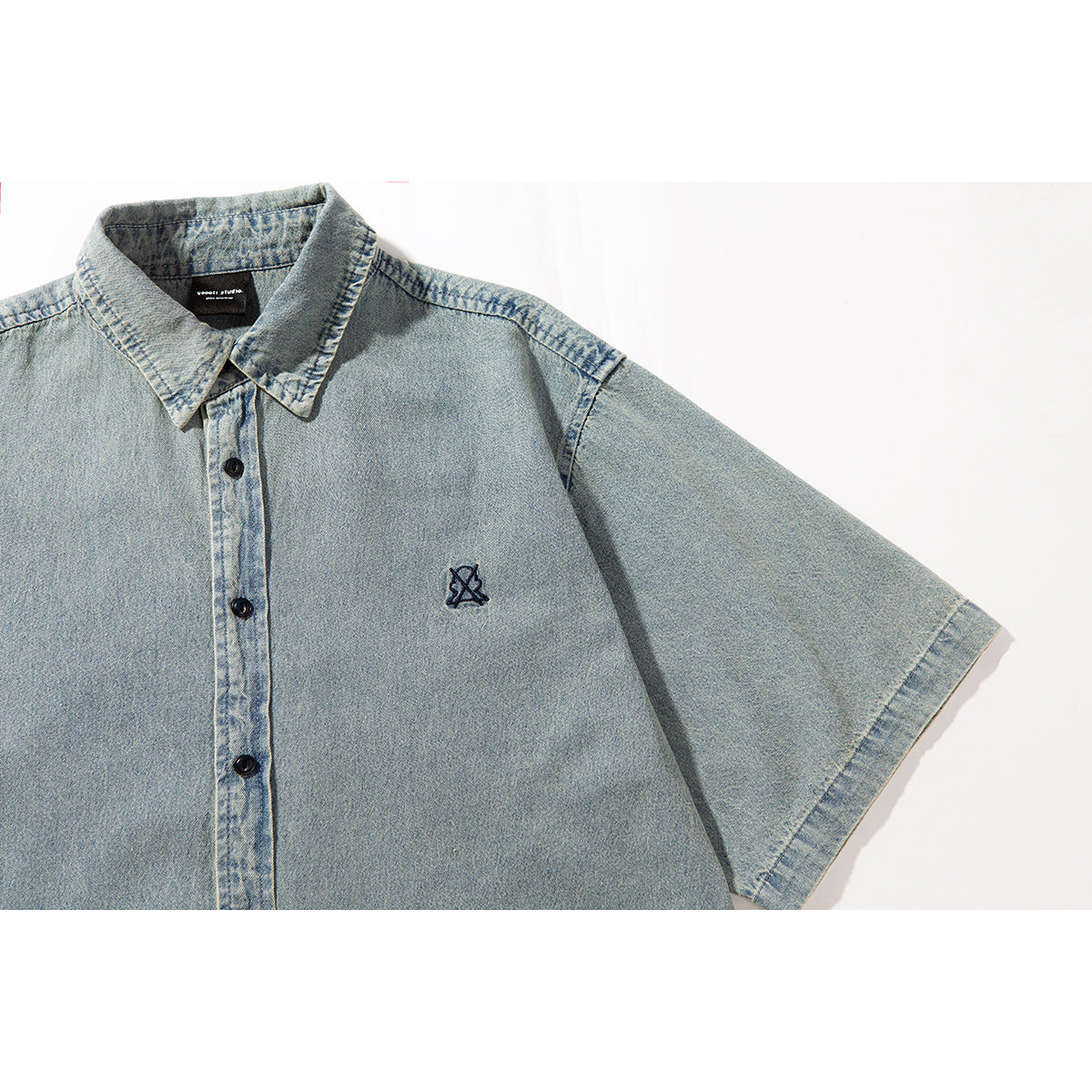 Close-up of a light blue short-sleeve denim shirt with black buttons and a small embroidered chest emblem.