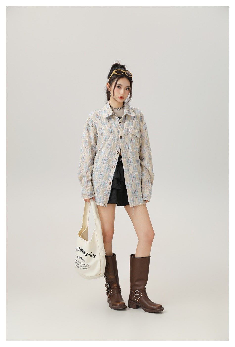 Model wearing buttoned beige plaid oversized shirt with black mini skirt, brown boots and a canvas tote bag
