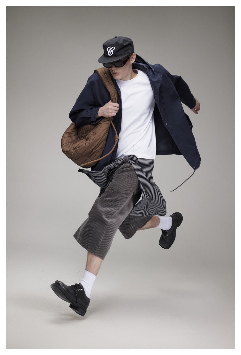 Model in motion wearing an open navy hooded jacket over a white tee, with cap, sunglasses, striped top and corduroy shorts