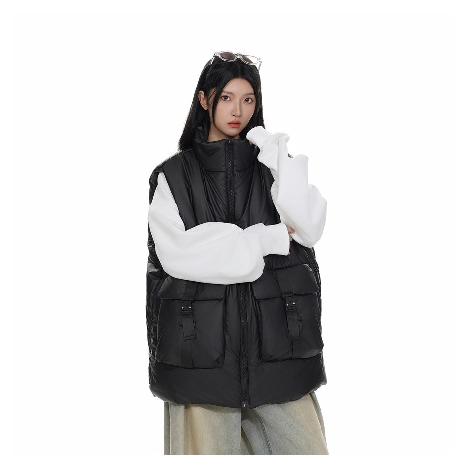 Model wearing a black high-collar padded vest with a front zipper and two large buckle-strap pockets over a white sweatshirt.