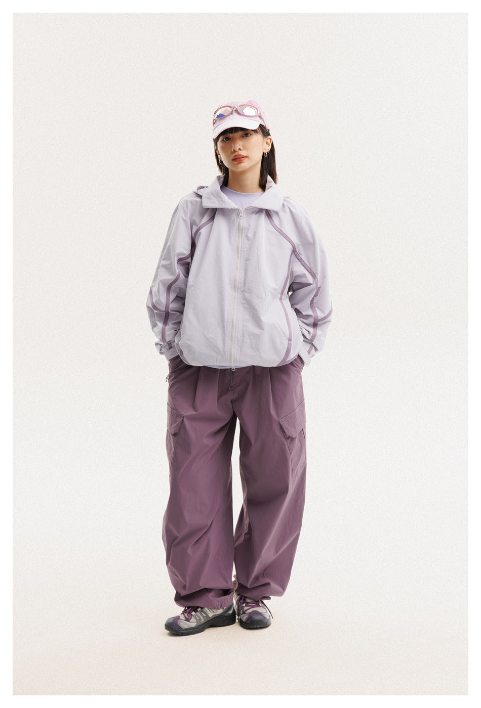 Front view of a woman wearing a light lavender zip jacket and plum cargo pants with a pink cap.