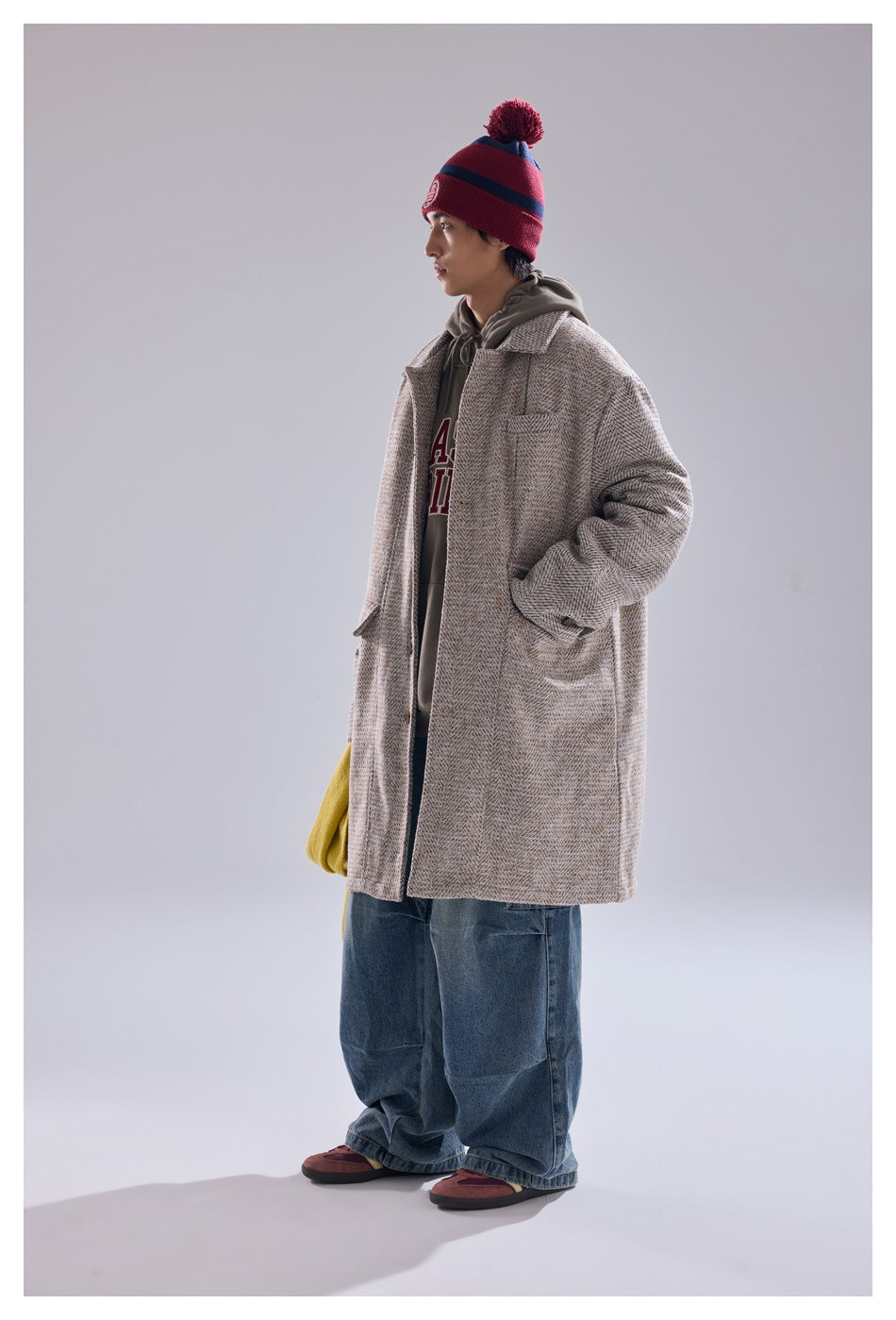 Person in khaki herringbone mid-length coat over a gray hoodie, wide jeans and a red striped beanie, side view