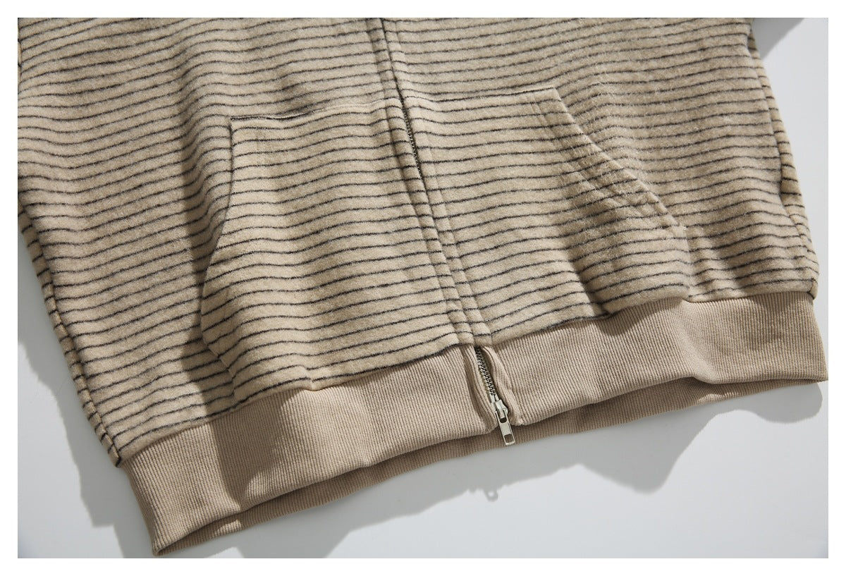 Close-up of a beige jacket with thin dark stripes, a ribbed hem, and the bottom of its front zipper.