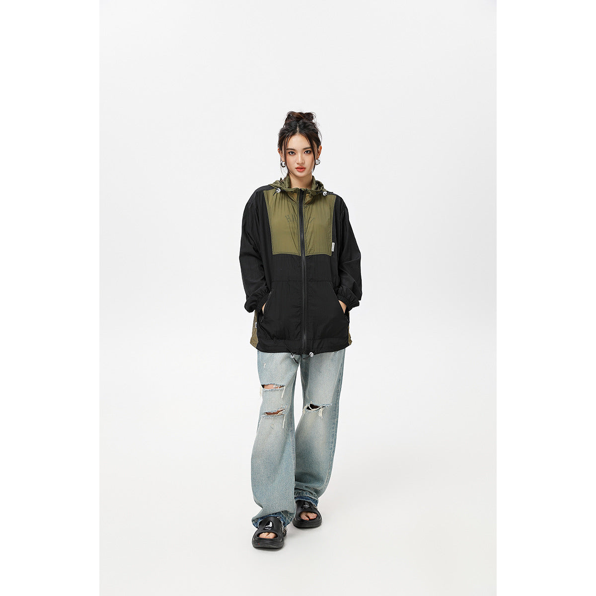 Front view of a model wearing a zipped black and olive hooded windbreaker with loose distressed jeans.