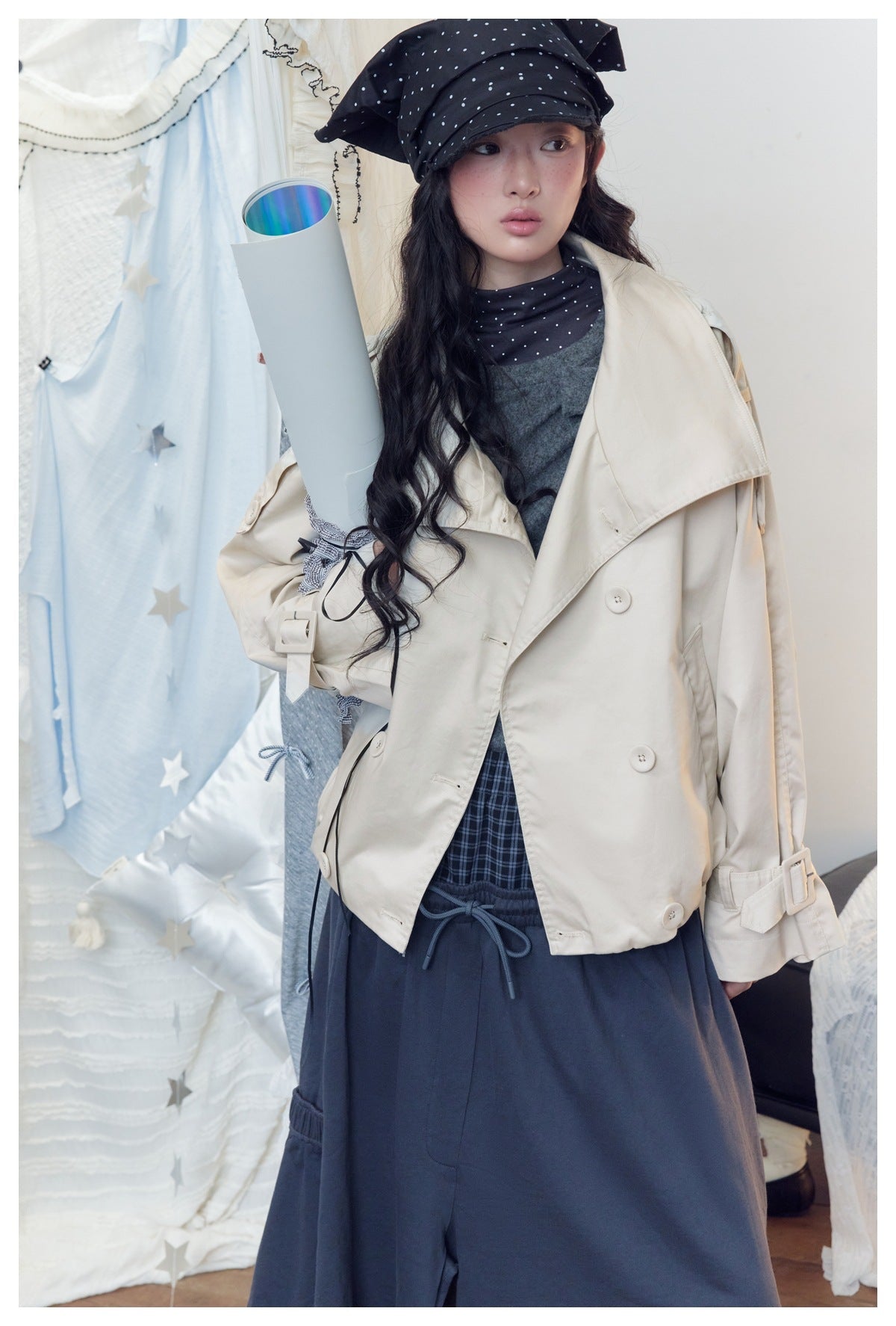 Model wearing a cream oversized double-breasted jacket over a gray top and navy bottoms, with a black polka-dot headscarf.