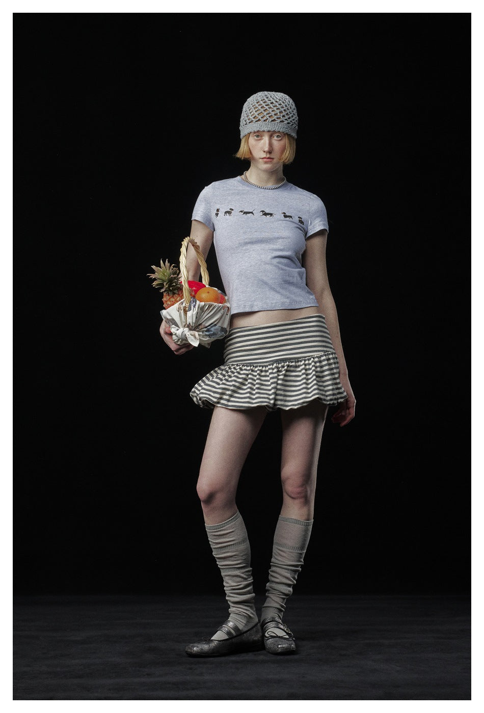 Full-length model in a pale gray dog-print T-shirt, striped ruffle skirt, gray leg warmers and open-knit beanie.