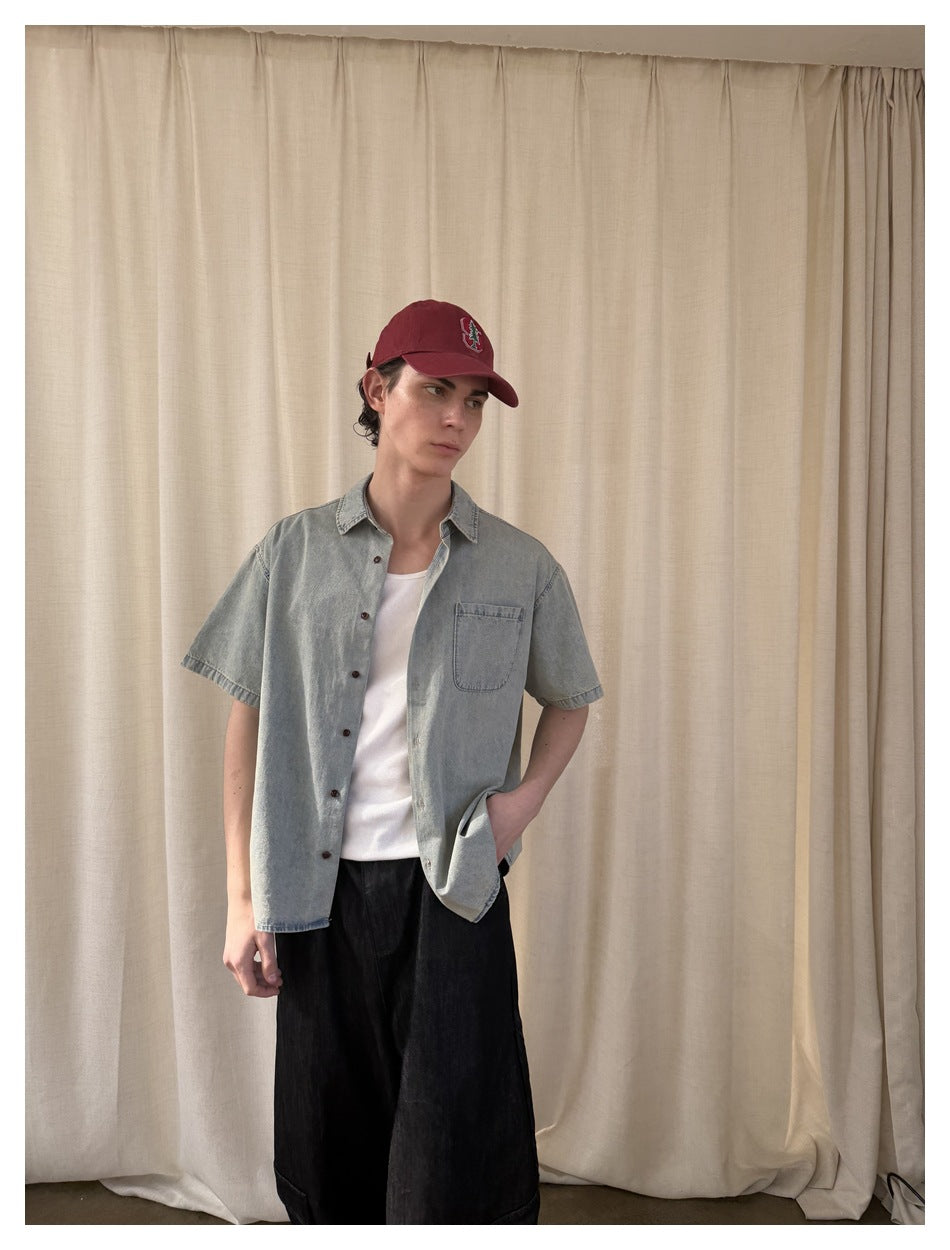 Model in an open light blue short-sleeve shirt over a white top and wide black trousers, posing at an angle.