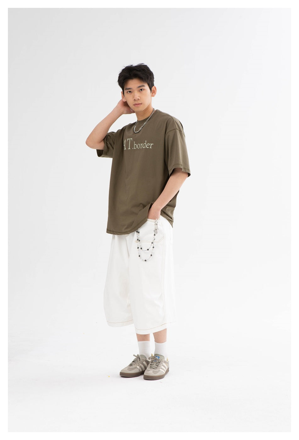 Model wearing a loose olive T-shirt with white lettering and wide white knee-length shorts with a chain detail.