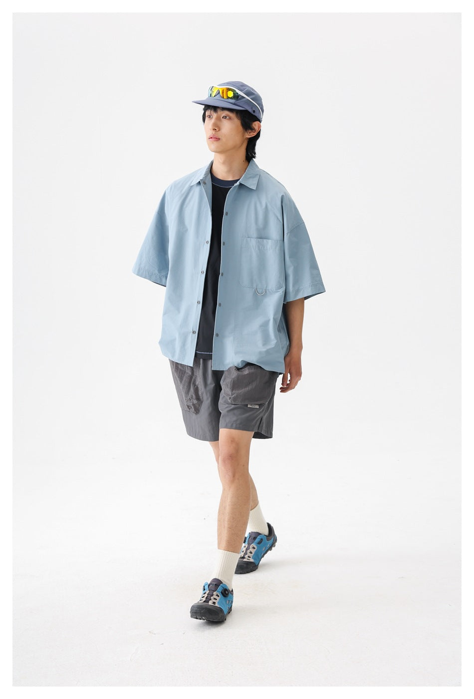 Model walking in a pale blue oversized short-sleeve button-up shirt with a chest pocket, viewed from the front.