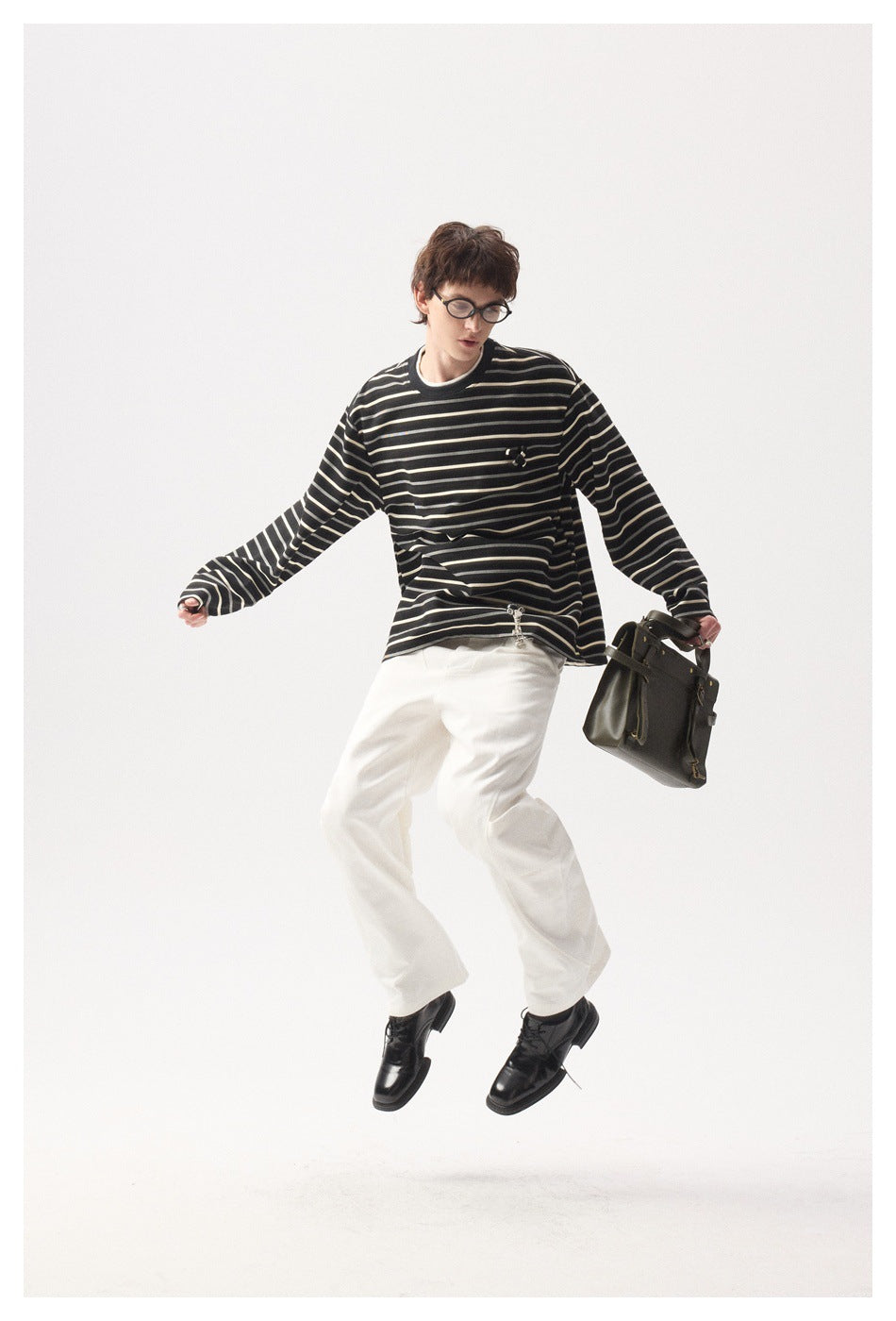 Model in a black striped long-sleeve shirt and white trousers, shown mid-jump with a dark bag.