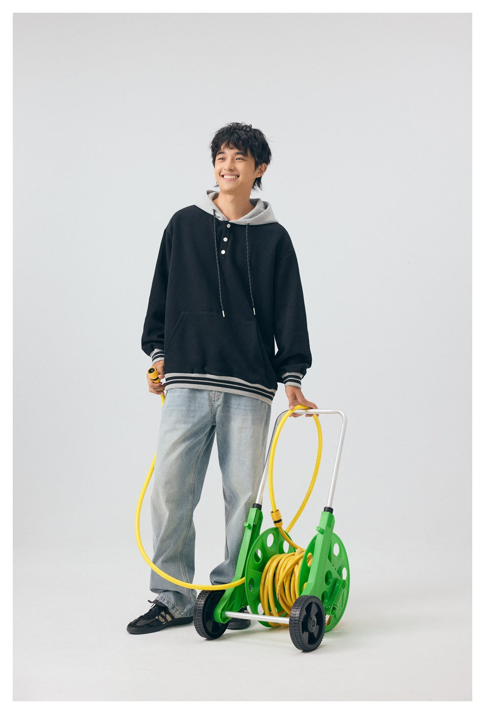 Model in black hoodie with gray hood and striped trim and light jeans, standing with a garden hose reel