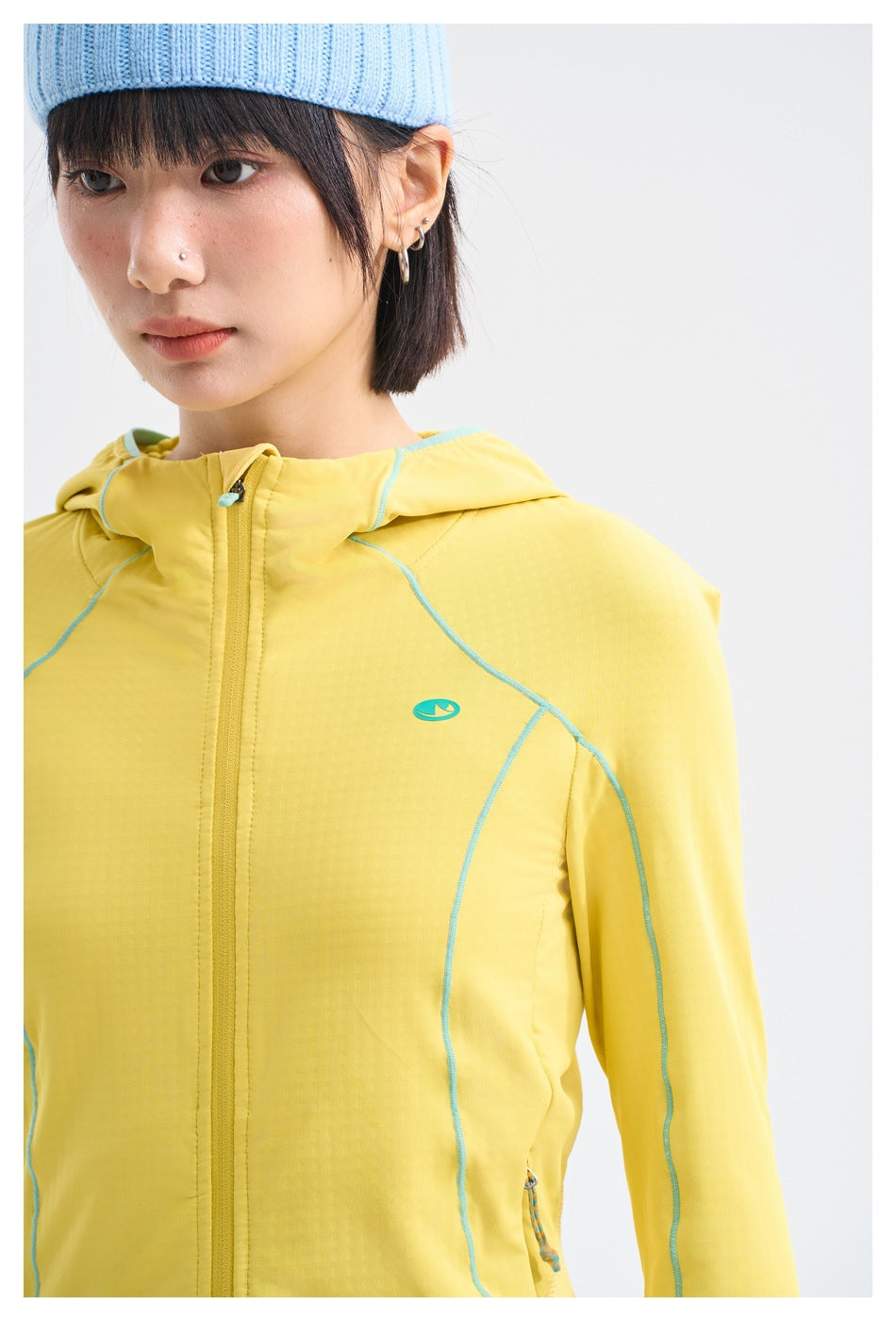 Close-up of a yellow hooded zip jacket with turquoise piping and a small chest emblem, worn by a model.