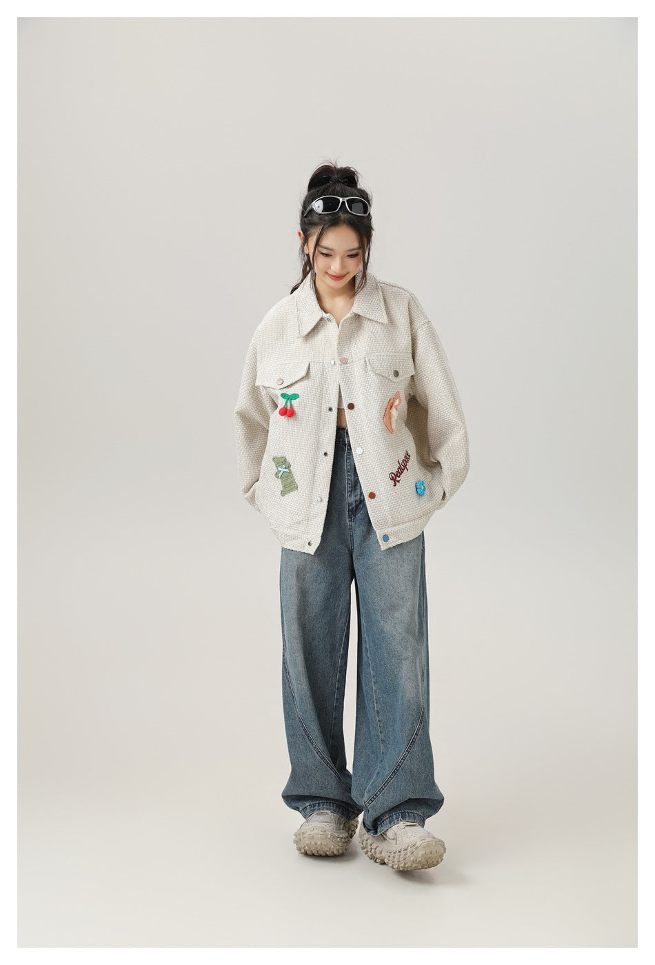 Model wearing the light khaki appliqué jacket open over a cropped top with wide-leg blue jeans and chunky sneakers