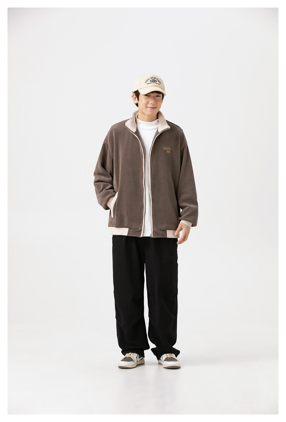 Full-length model wearing a taupe ribbed jacket with cream trim, black trousers and a cap.
