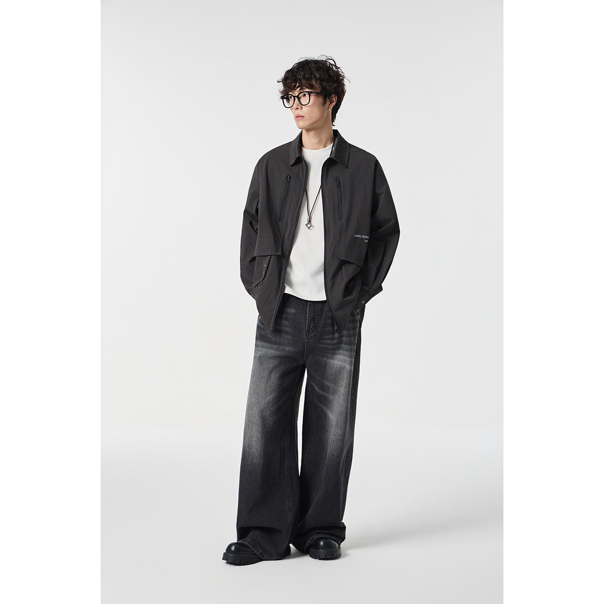 Model wearing a black utility jacket over a white shirt with loose dark gray jeans.