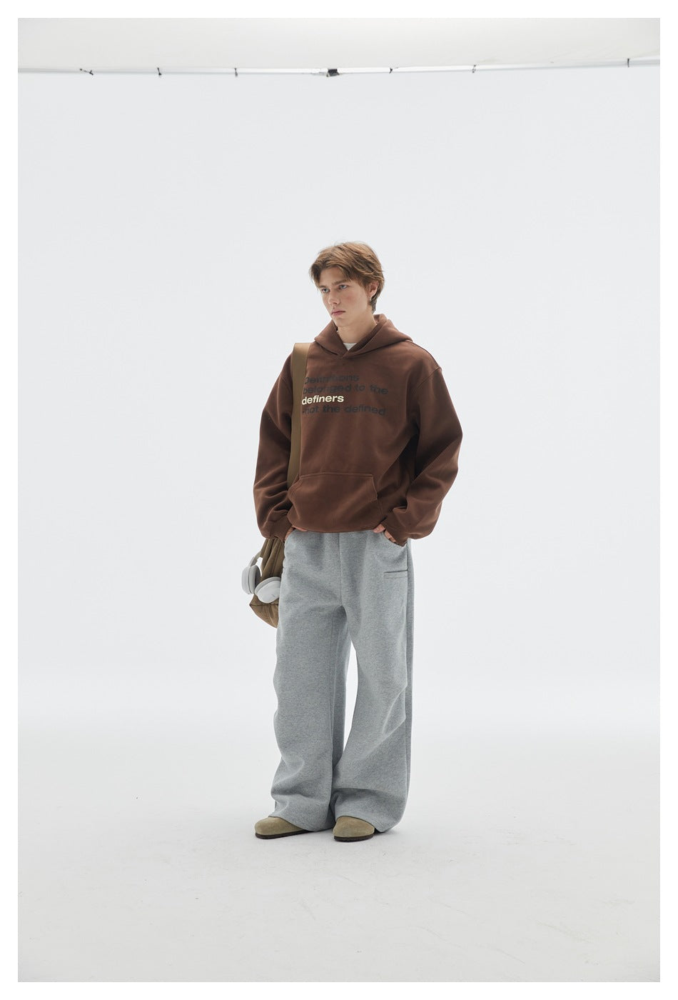 Full-length front view of a model in a brown text-print hoodie and wide gray pants, with a shoulder bag and headphones.