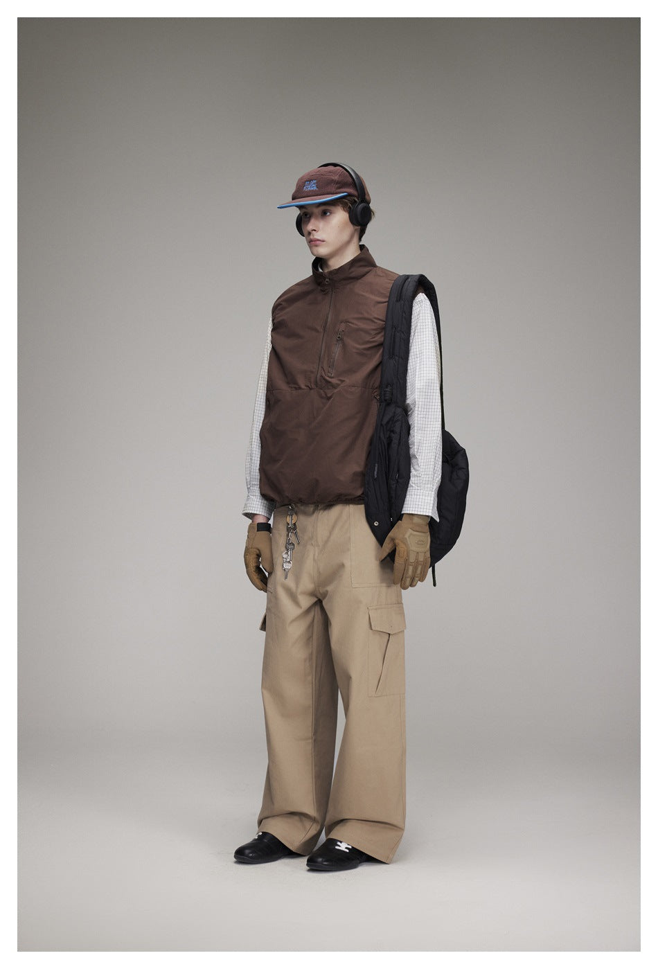 Model wearing a brown half-zip vest over a checked shirt with khaki cargo pants, cap, headphones and gloves