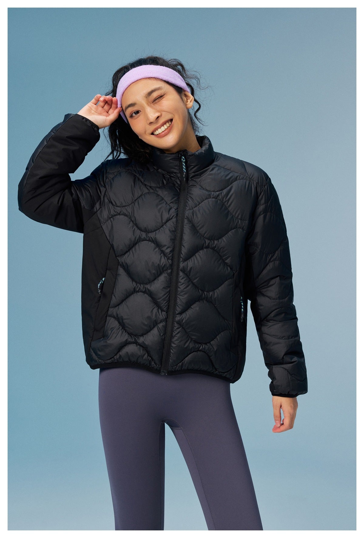 Woman modeling a black wave-quilted zip jacket with a high collar and purple leggings, shown from the front.