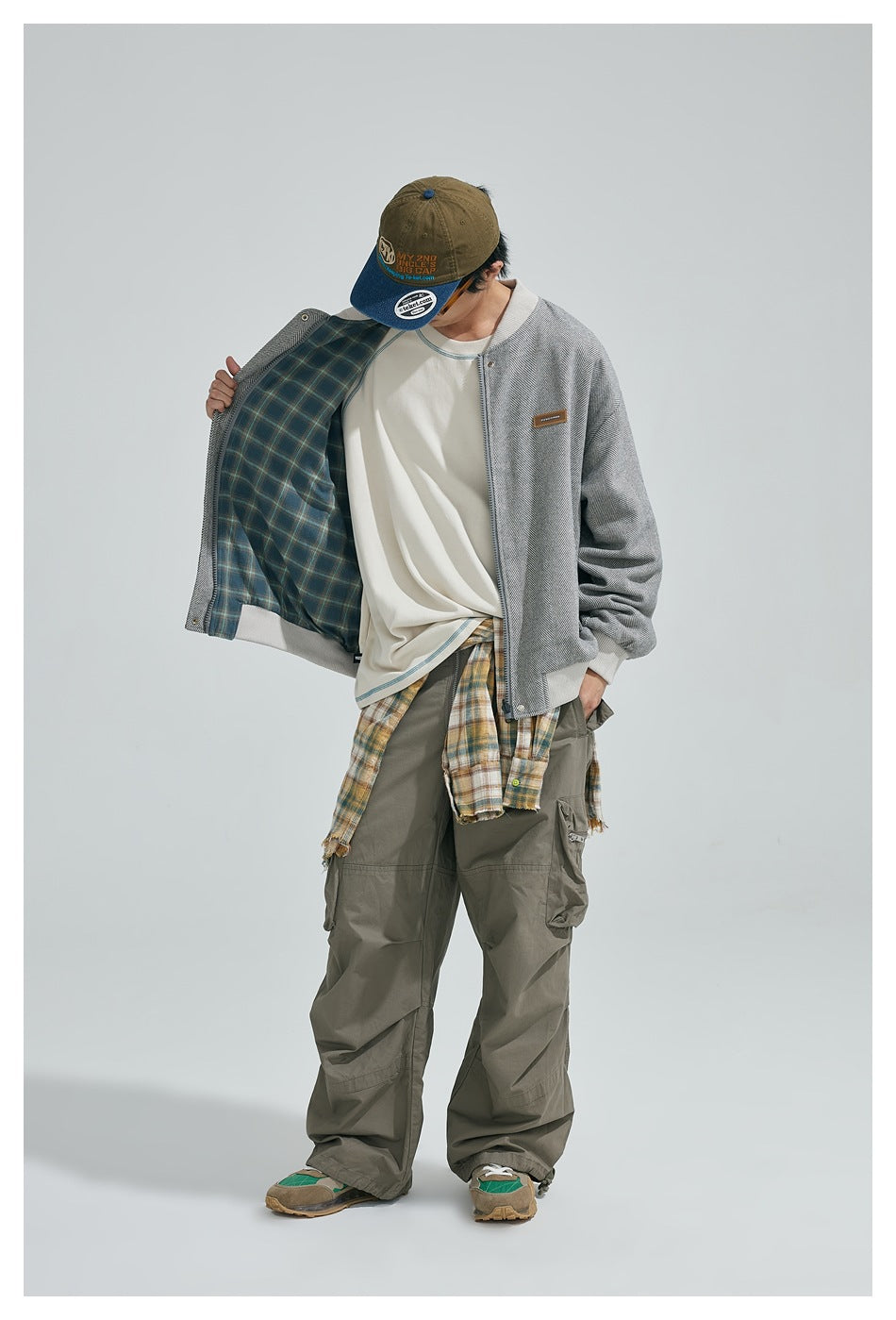 Model holding jacket open to show the blue-green plaid reverse side, worn with cream tee and olive cargo pants