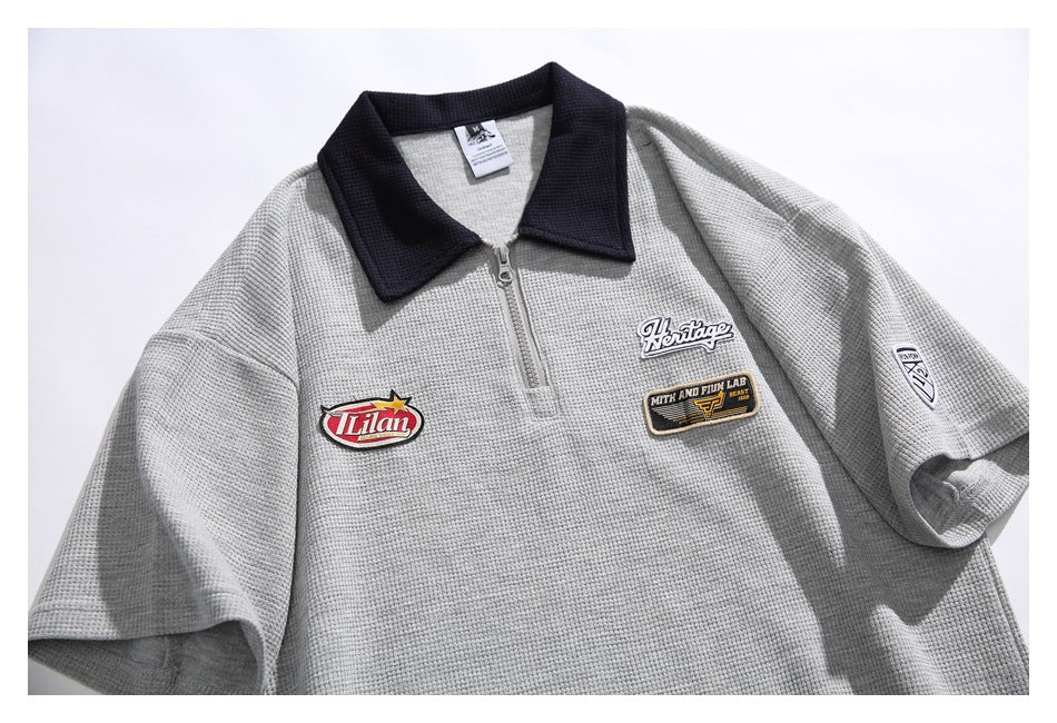 Front view of a light gray textured short-sleeve pullover with a dark collar, quarter zipper, and embroidered patches.