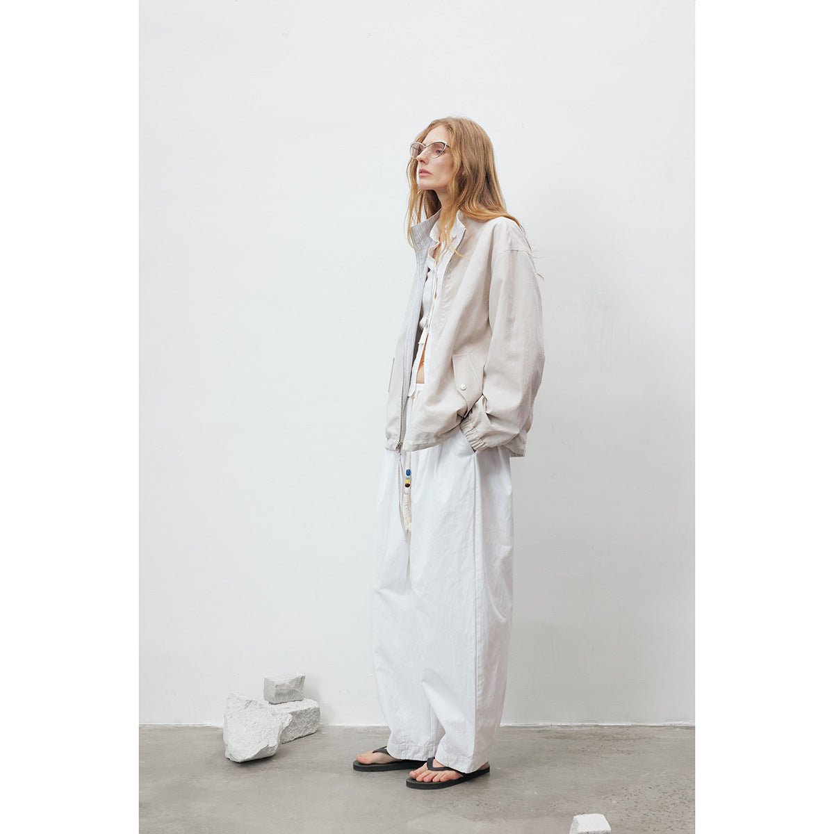 Model in a pale beige zip jacket and loose white trousers, shown in profile wearing glasses and sandals.