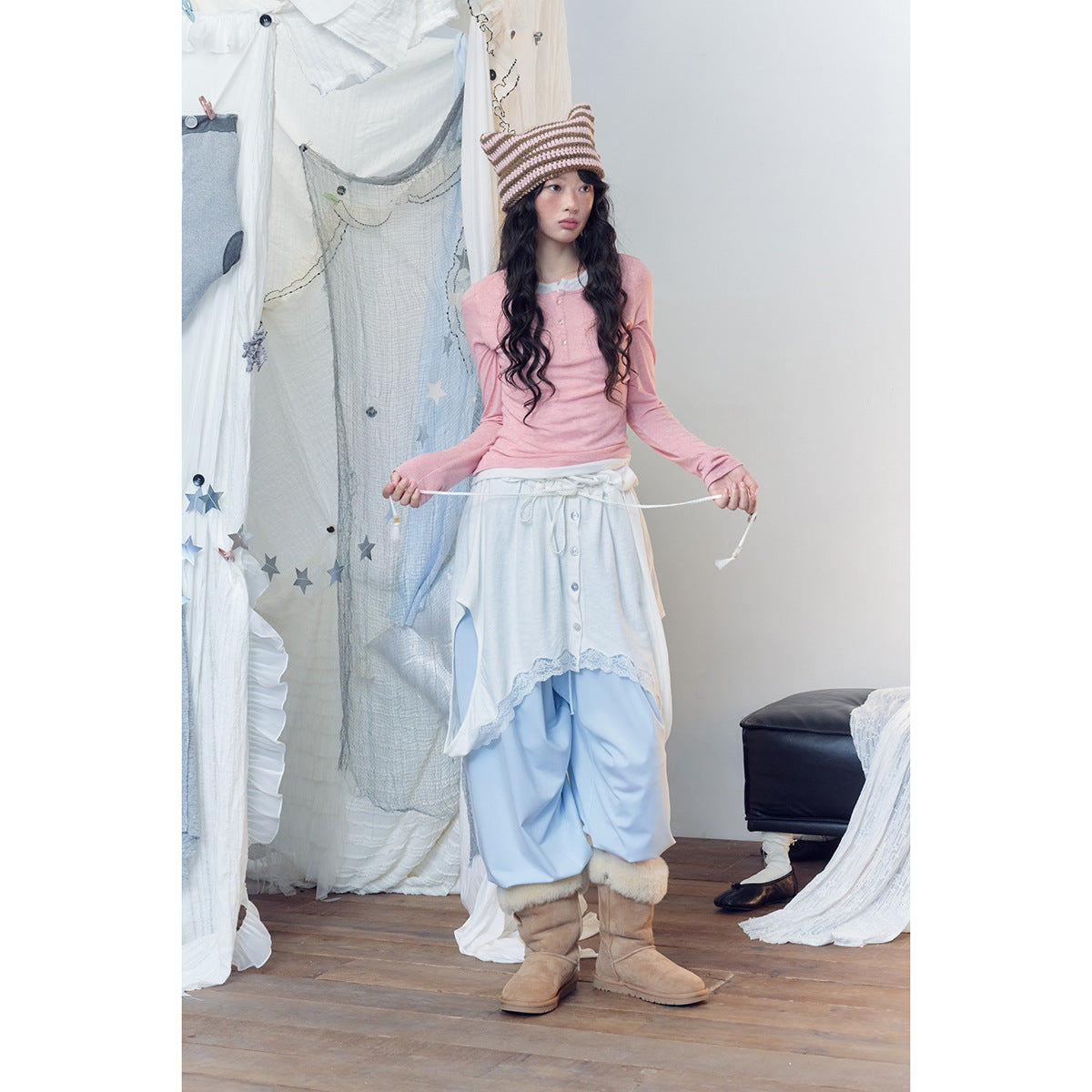 Model wearing a pink long-sleeve henley, white lace-trimmed wrap skirt, pale blue trousers and tan boots.