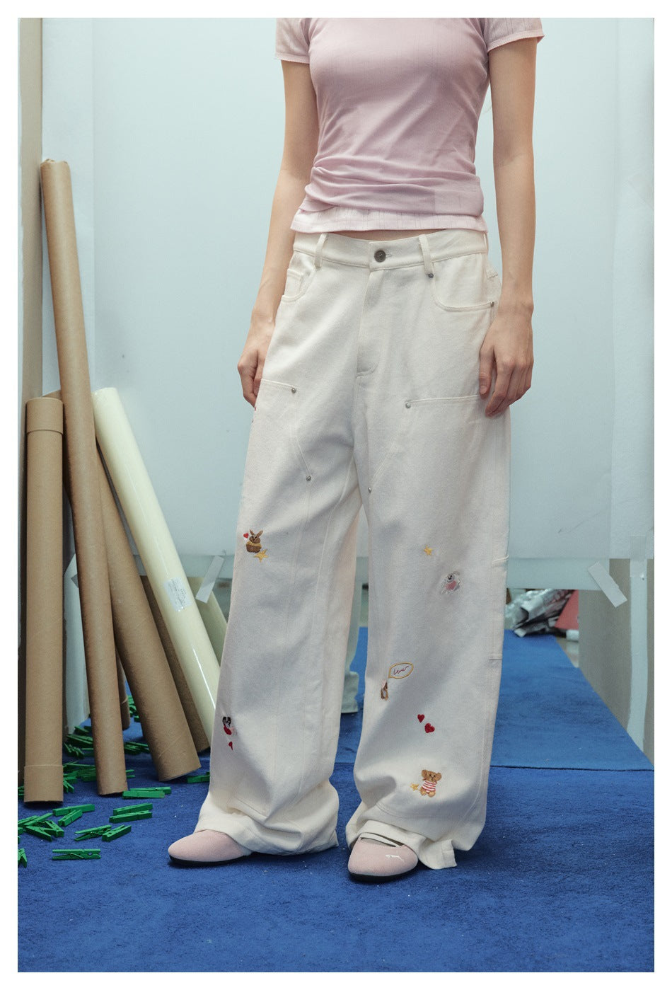 Front view of a model wearing cream wide-leg trousers with utility pockets and small embroidered motifs.