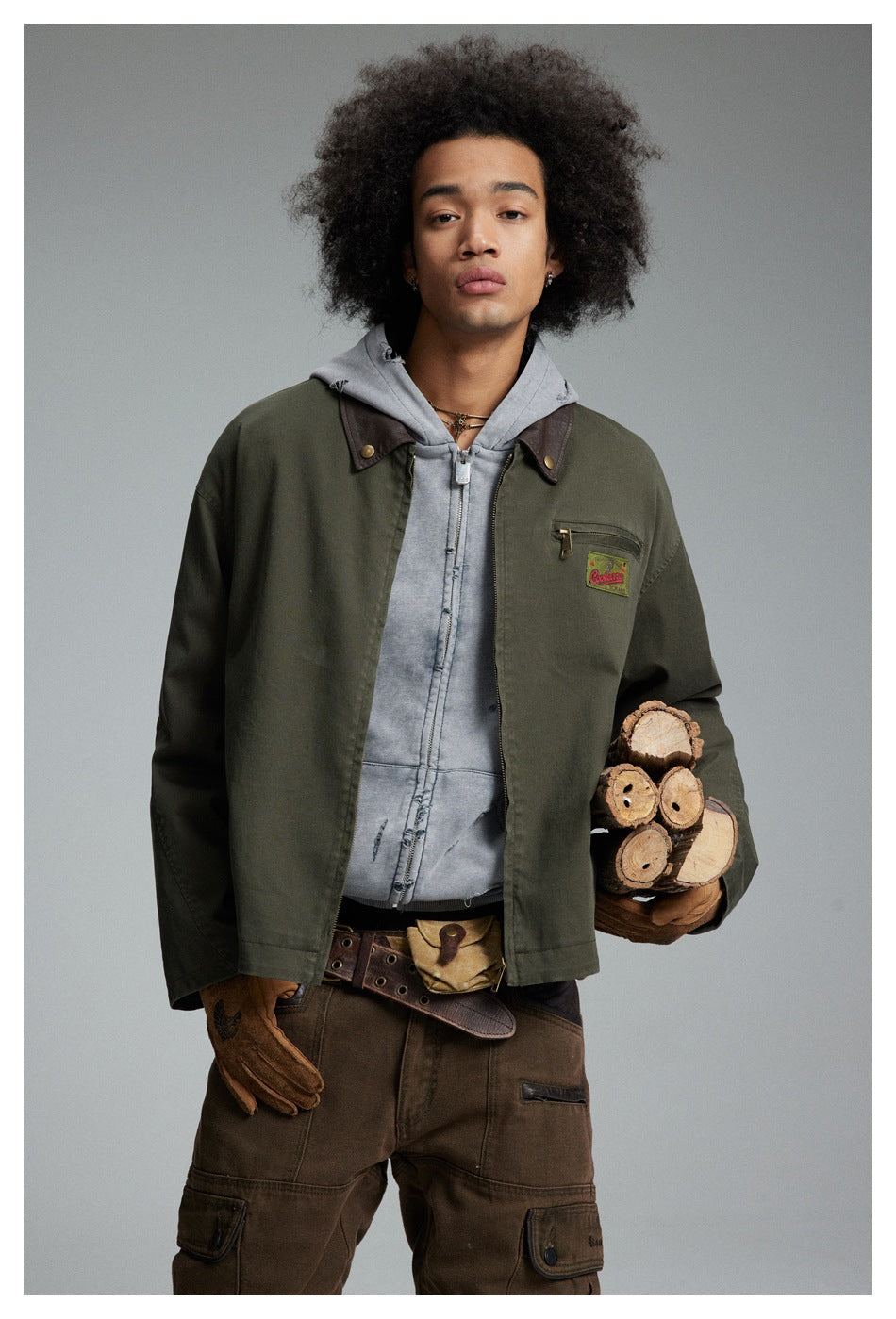 Front view of a model wearing an olive jacket over a gray hoodie, holding a bundle of cut wood.