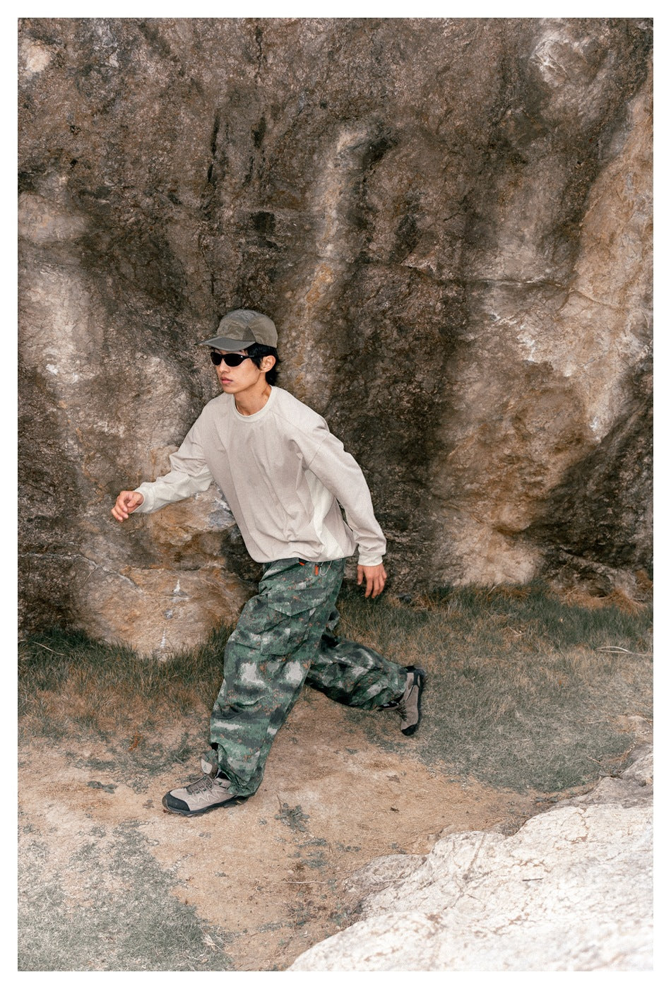 Model in a cream long-sleeve shirt and green camouflage cargo pants stepping across rocky ground.