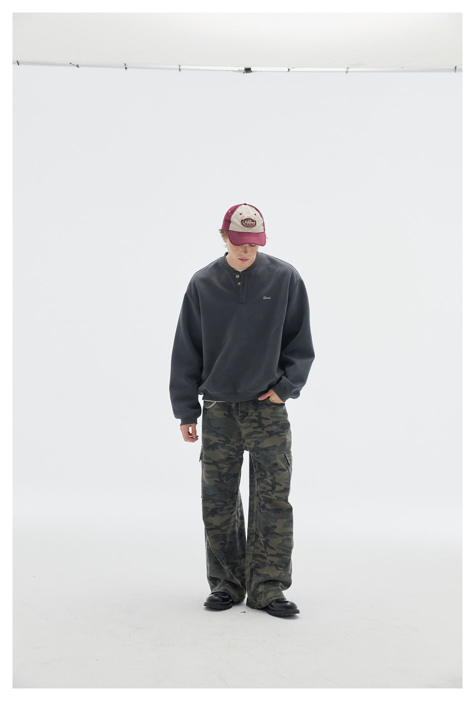 Full-length model wearing a charcoal henley sweatshirt, camouflage cargo pants and a burgundy-and-cream cap.