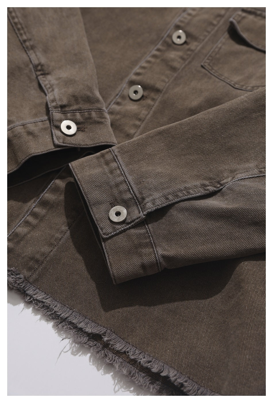 Brown-gray shirt close-up showing snap-button cuffs, a chest pocket, stitching, and a frayed hem.