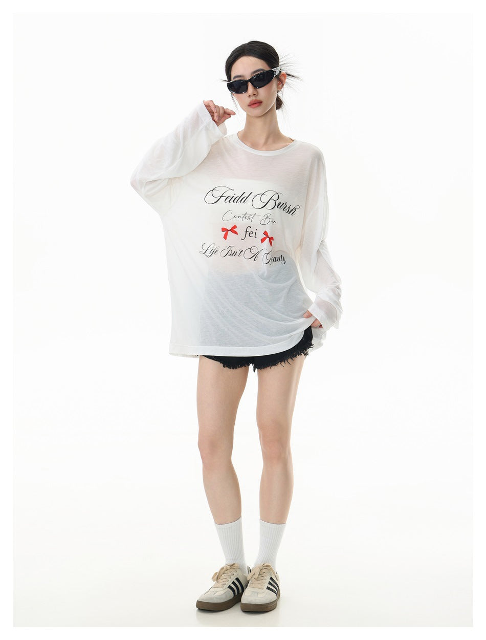 Model wearing an oversized semi-sheer white long-sleeve top with script print and red bows, black shorts and sneakers