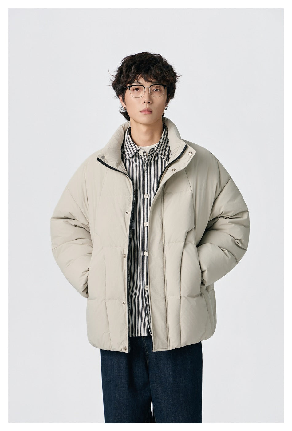 Man wearing an open beige puffer jacket with stand collar over a striped button-up shirt, hands in pockets